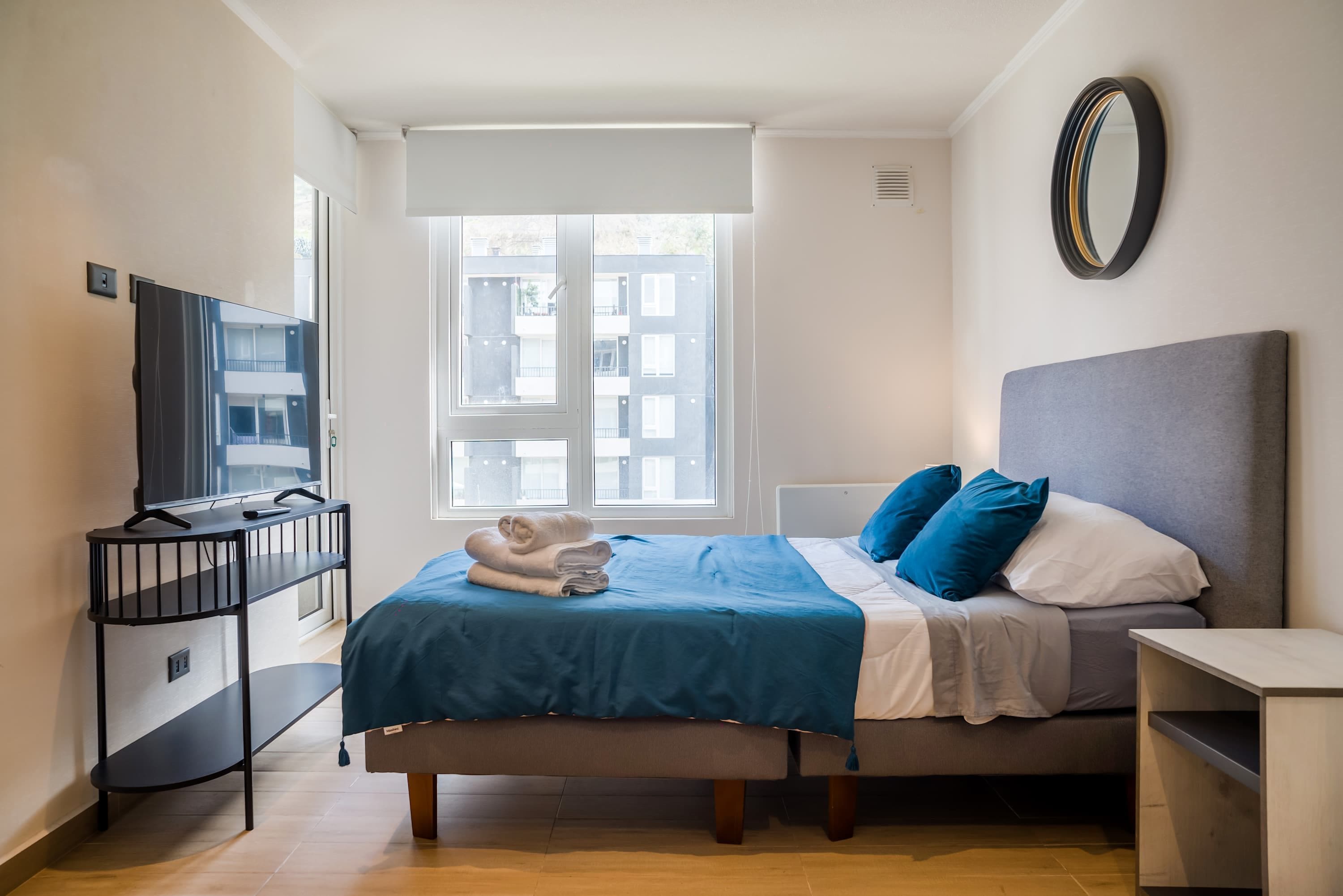 Habitación de hotel con cama, televisor en un mueble bajo, ventana con persianas y espejo en la pared.