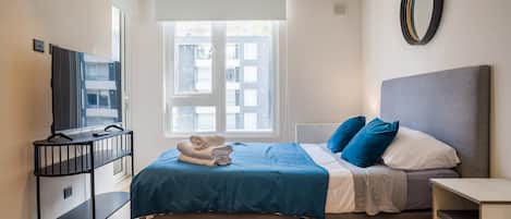 Habitación de hotel con cama, televisor en un mueble bajo, ventana con persianas y espejo en la pared.