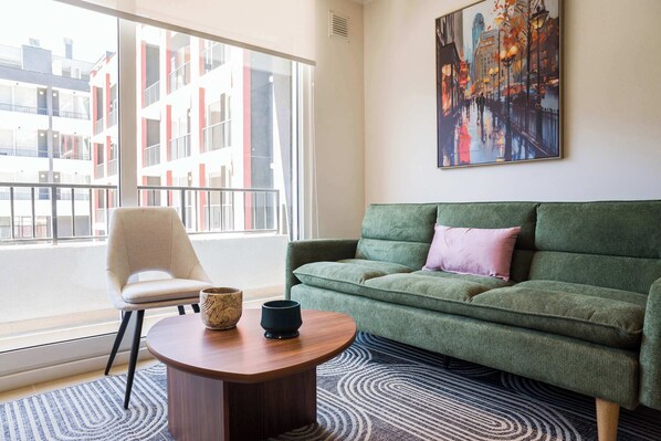 Una sala de estar moderna con un sofá verde, un sillón beige, una mesa de centro redonda de madera y un ventanal con vistas a la ciudad.