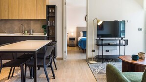 Une cuisine moderne avec des meubles en bois, une table à manger et des chaises. On aperçoit une chambre à coucher à travers une porte ouverte, avec un lit et une table de chevet. Un téléviseur est fixé au mur dans le salon.