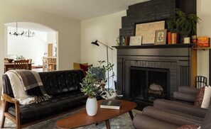 Una sala de estar con un sofá de cuero negro, una mesa de centro de madera, una chimenea y un comedor al fondo.