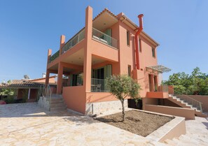 A two-story building with a balcony, a staircase, and a small tree in the courtyard. -  COZY NEWLY BUILD BY THE SEA APARTMENT IDEAL FOR FAMILIES (Katakolo)