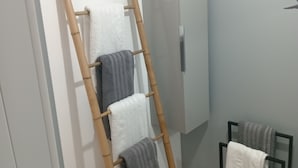 Une salle de bain avec un porte-serviettes en bambou où sont rangées des serviettes, un meuble blanc et une petite étagère noire sur laquelle repose une serviette.