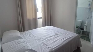 A hotel room with a bed, curtains, a bathroom visible through an open door, and a small window. - Apartment in Summer Park (Florianópolis)