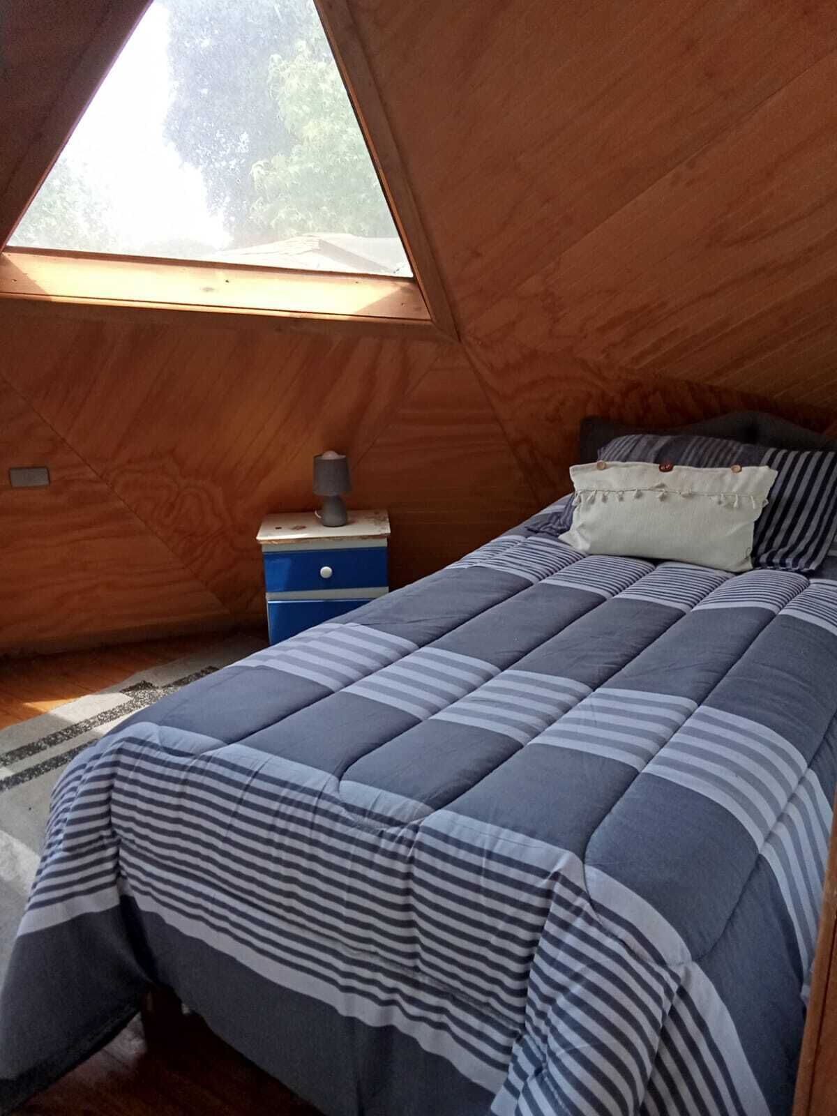 A bedroom with a sloped ceiling, a bed with striped bedding, a nightstand with a lamp, and a window with a view of trees.