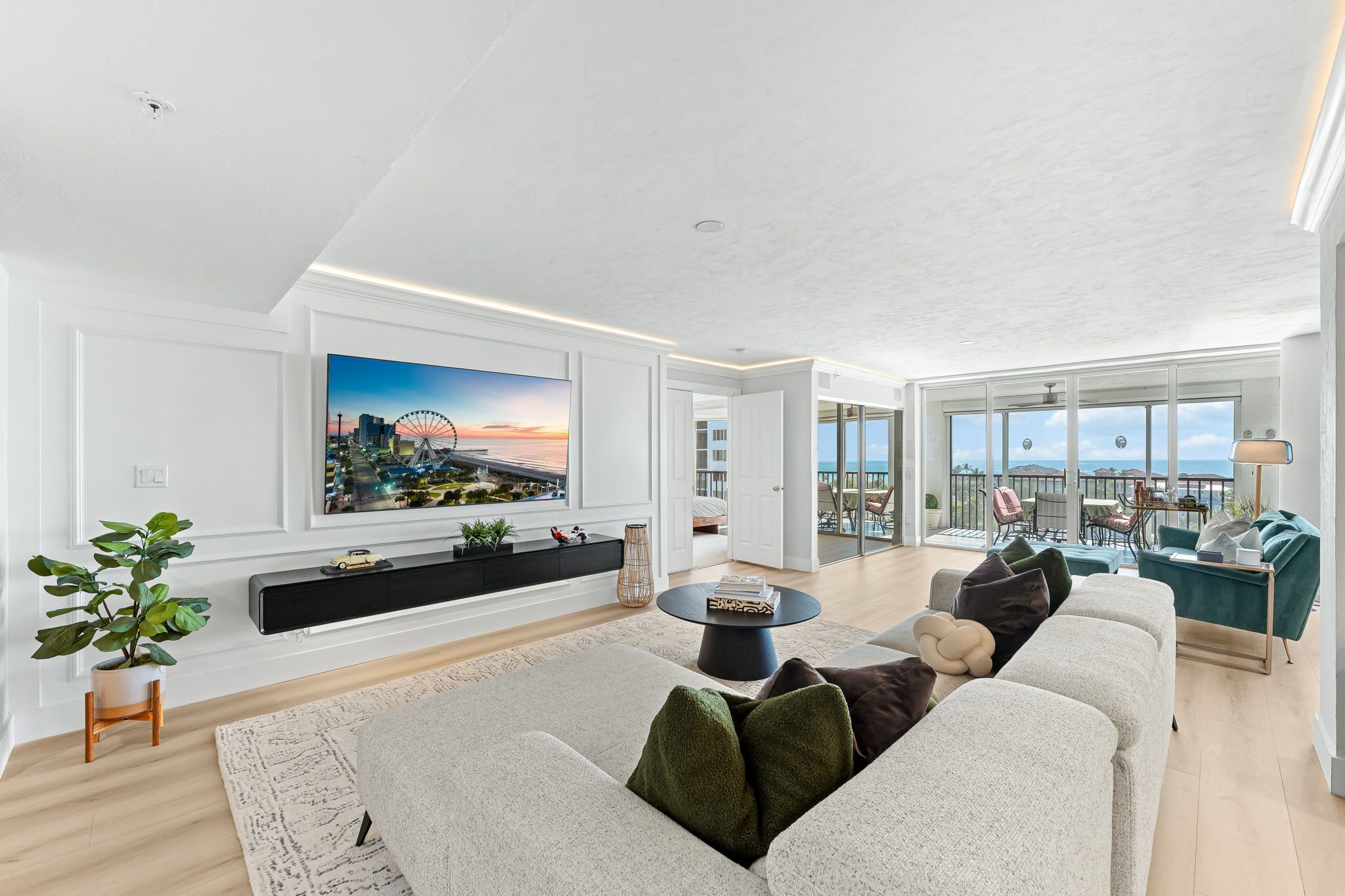 A spacious living room with a large flat-screen TV, a comfortable sofa, a coffee table, and a balcony with a view of the city.