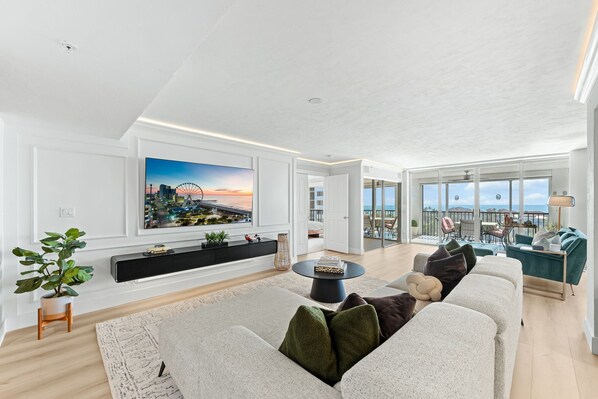 A spacious living room with a large flat-screen TV, a comfortable sofa, a coffee table, and a balcony with a view of the city. - Gorgeous Ocean Views, Pool, Amenities Galore, Condo Living at it Finest! (Fort Myers Beach)