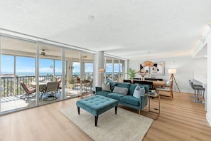 A spacious living area with a large window overlooking the ocean, a teal sofa, and a dining area with a view. - Gorgeous Ocean Views, Pool, Amenities Galore, Condo Living at it Finest! (Fort Myers Beach)