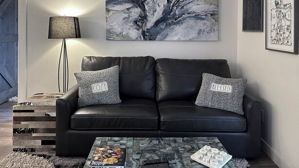 A modern living room with a black leather sofa, two grey patterned pillows, a glass coffee table with remote controls and a magazine, a floor lamp, and abstract wall art. - Red Pine Retreat at Canyons Village (Park City)