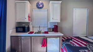 A compact kitchen with a microwave, toaster, and coffee maker. There is a sink with a faucet, and a clock on the wall. A small refrigerator is visible, and a bed with a striped blanket is in the background. - All-american Escape, Riverfront! WB3 (Parker Dam)
