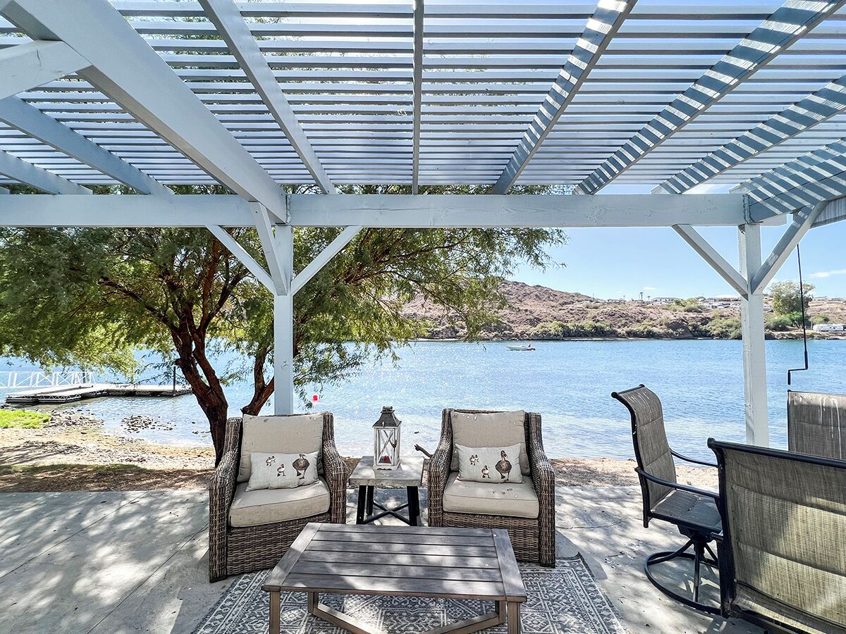 A covered outdoor seating area with wicker chairs and a table, overlooking a body of water.