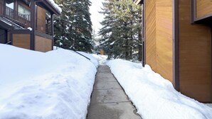 A snow-covered pathway between buildings with wooden siding and metal railings. - Relaxing Canyons Condo Free Shuttle Ride To Lift (Park City)