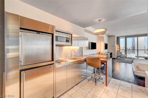 A modern kitchen with stainless steel appliances, a dining area with a bar stool, and a large window offering a city view. - High Floor Mountain-views Studio Balcony Access (Las Vegas)