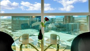 Una mesa con dos tazas blancas y un jarrón negro con una sola rosa roja, con vistas a un paisaje urbano a través de amplias ventanas.