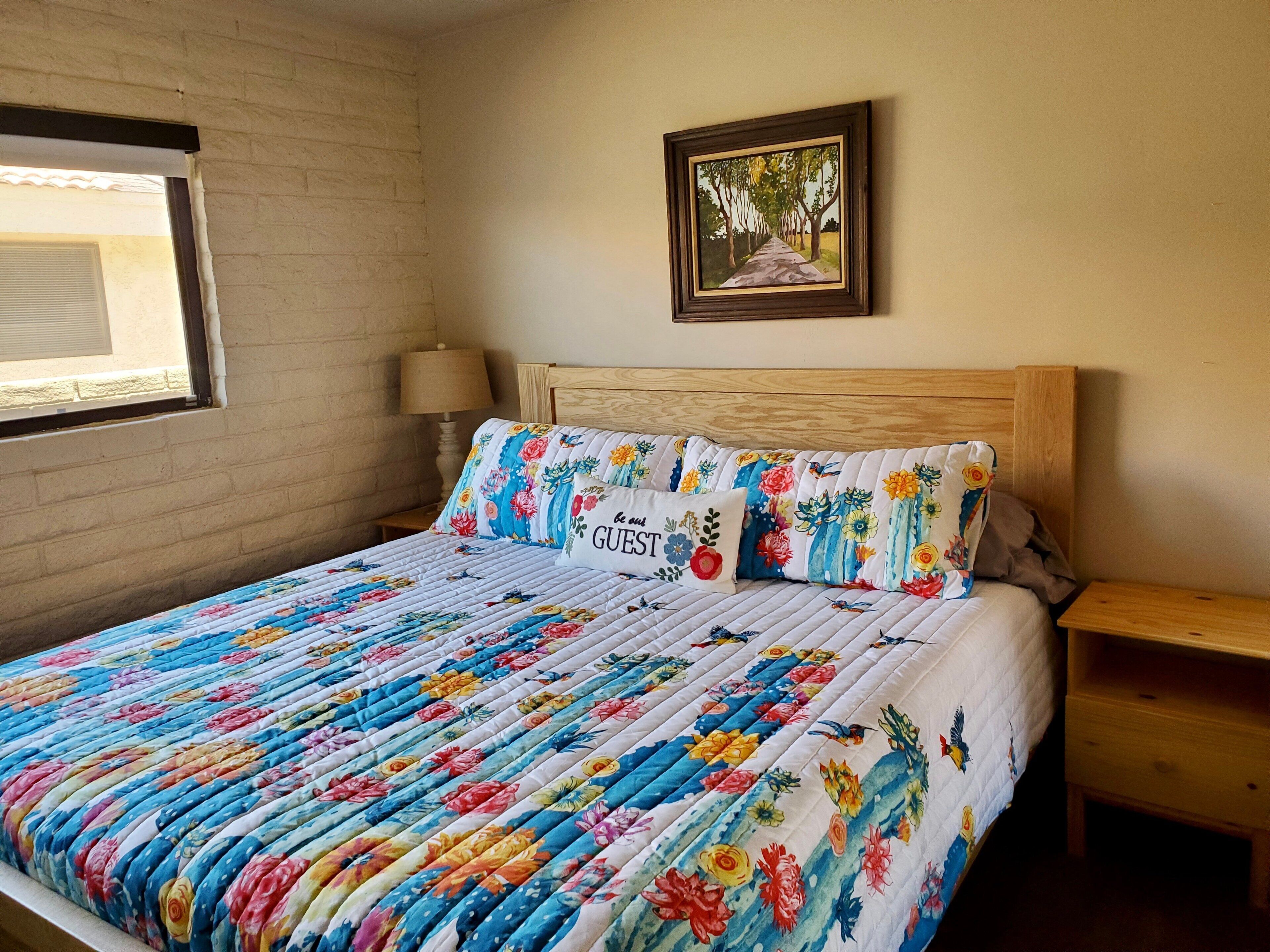 A bedroom with a floral quilted bedspread, a wooden headboard, a bedside table with a lamp, and a framed picture on the wall.