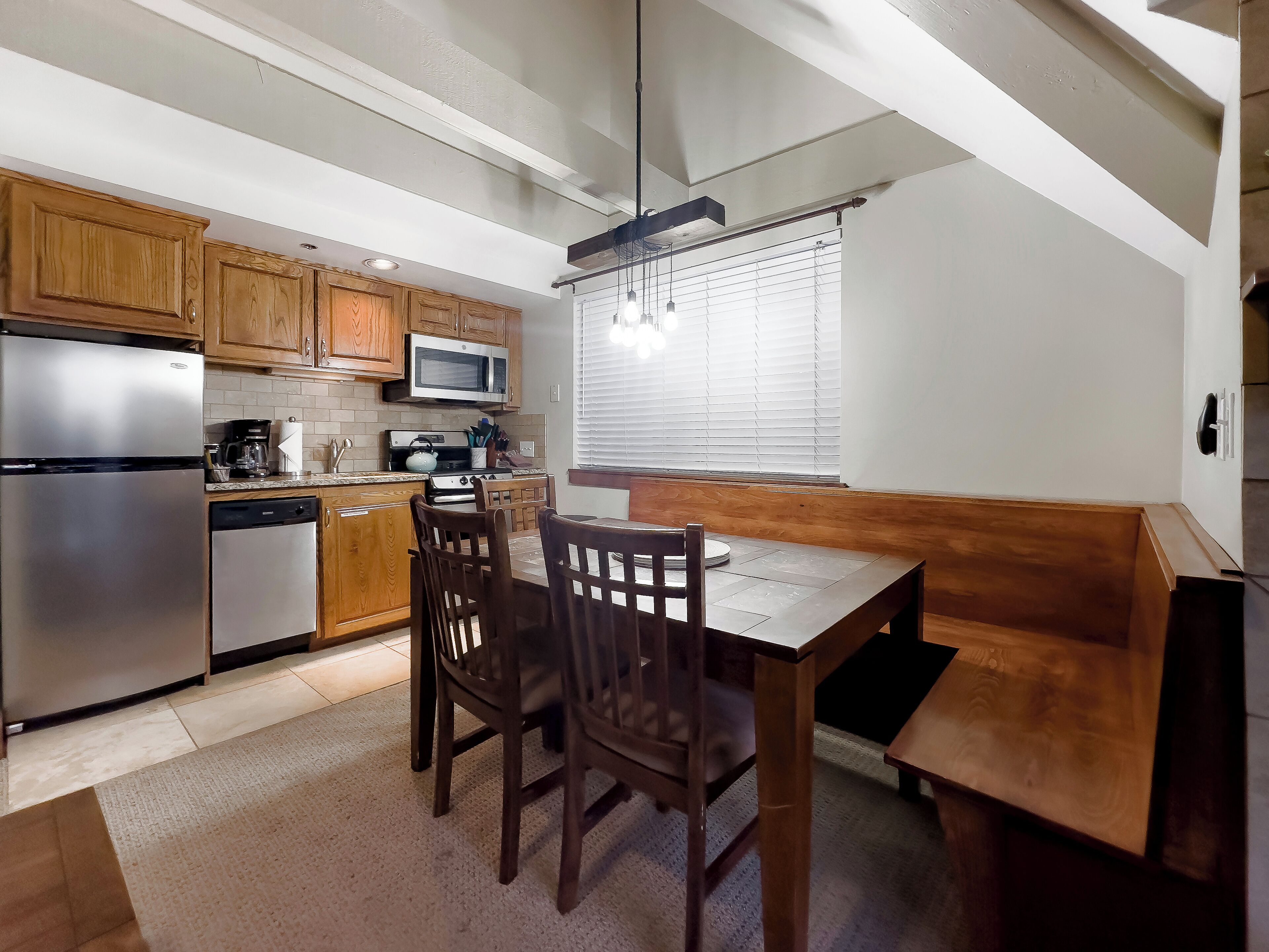 Μια μικρή, λειτουργική κουζίνα με ξύλινα ντουλάπια, ένα ψυγείο από ανοξείδωτο ατσάλι, ένα φούρνο μικροκυμάτων και μια κουζίνα. Υπάρχει επίσης ένα τραπέζι φαγητού με καρέκλες και ένα παγκάκι.