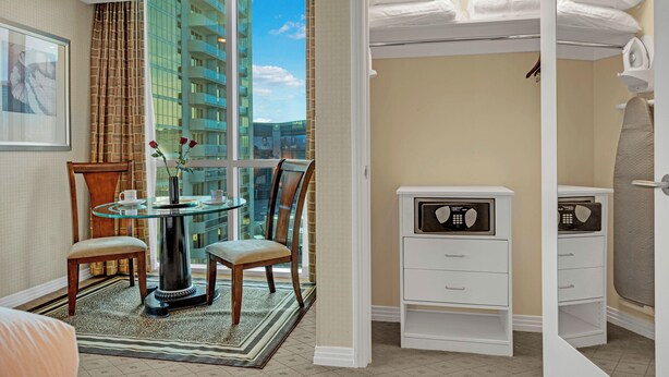 Habitación de hotel con una mesa de vidrio, sillas, una cama con cabecera blanca, un pequeño tocador blanco y vista a un edificio por la ventana.