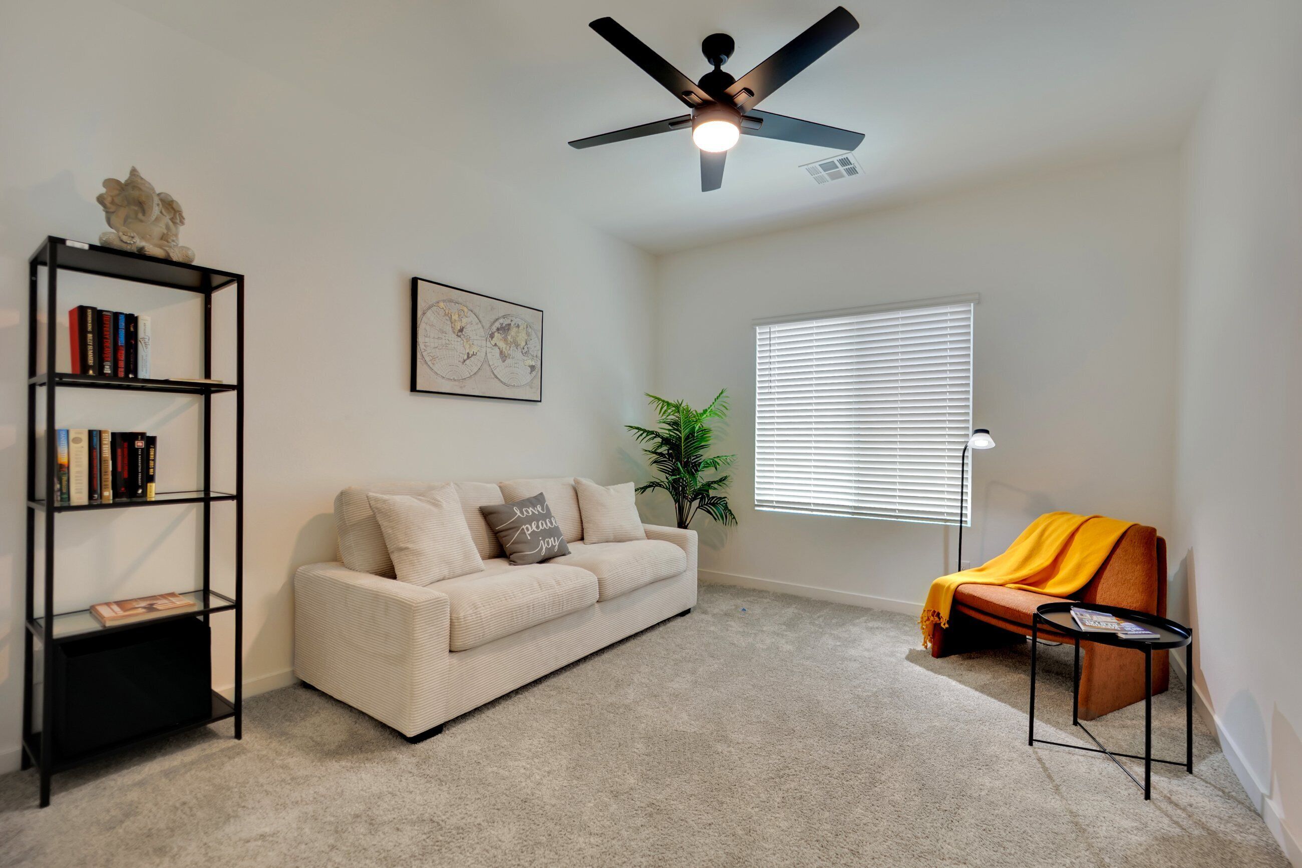 A living room with a beige sofa, a black bookshelf, a ceiling fan, a window with blinds, and a small table with a magazine.