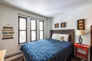 Une chambre à coucher avec un lit, une table de chevet et une fenêtre avec des stores. On y trouve des décorations murales et une lampe sur la table de chevet.