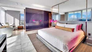 A modern hotel room with a large bed, a seating area with a chair and a small table, a large window with a view, and a glass partition. - Luxury 1BR Palms Strip View + Balcony Access 37322 (Las Vegas)