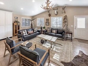 A spacious living room with a dark leather sofa, a wooden coffee table, and a large rug. There are three deer heads mounted on the wall, and a chandelier hangs from the ceiling. - Mountain Meadow Retreat Near Park City (Peoa)