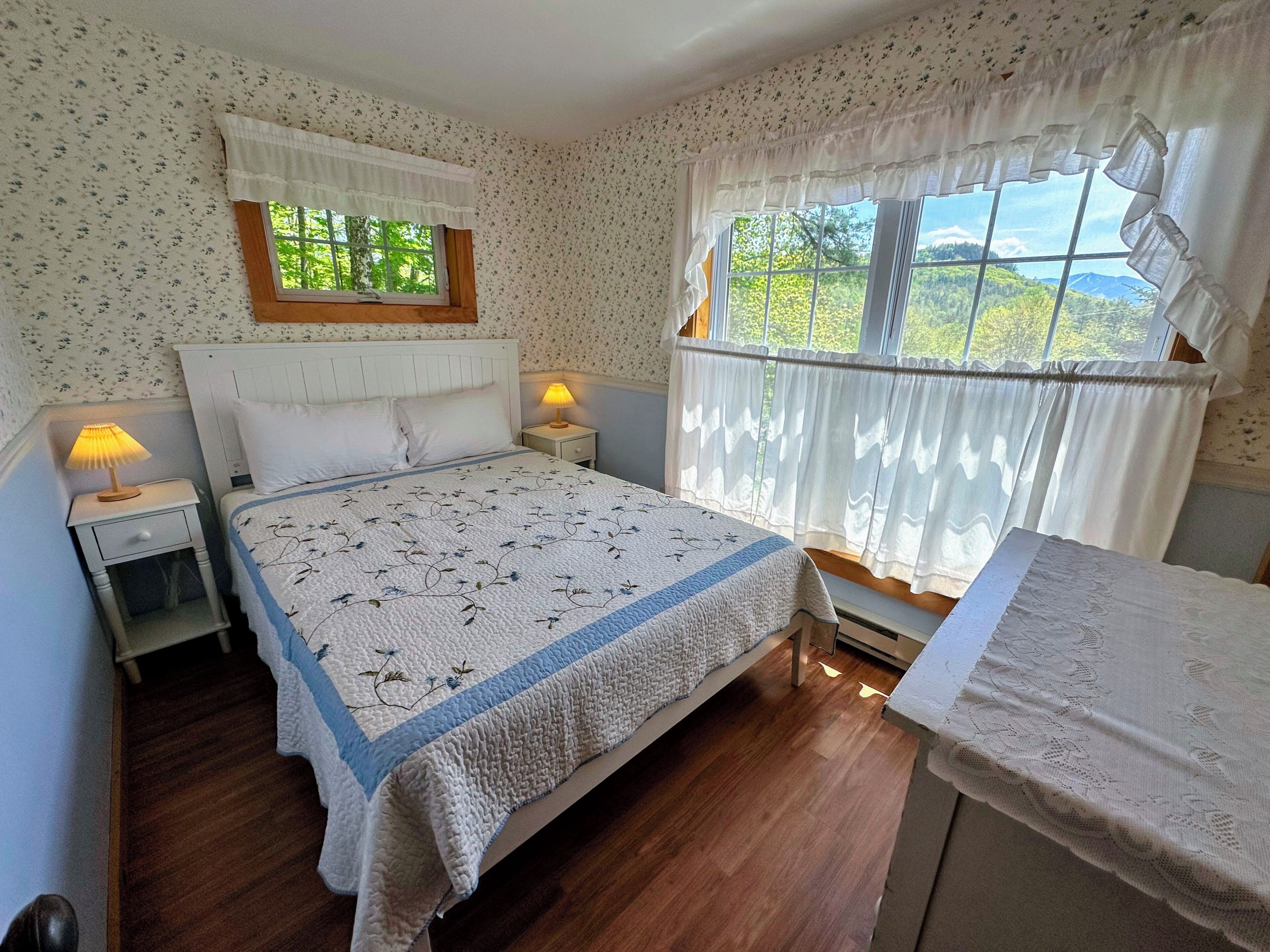 Une chambre à coucher avec un lit, deux tables de chevet, une fenêtre avec des rideaux et une vue sur la verdure à l’extérieur.