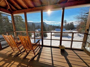 Een houten terras met schommelstoelen, een groot raam en uitzicht op een besneeerd landschap.