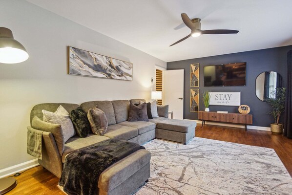 A modern living room with a sectional sofa, a ceiling fan, a flat-screen TV, and a decorative wall piece. - 425 Plantation Road SW, Smyrna (Smyrna)