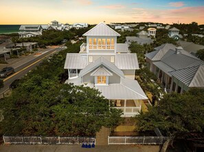 A large, multi-story house with a prominent central tower, surrounded by lush greenery and a coastal backdrop. - Four by the Shore (Santa Rosa Beach)