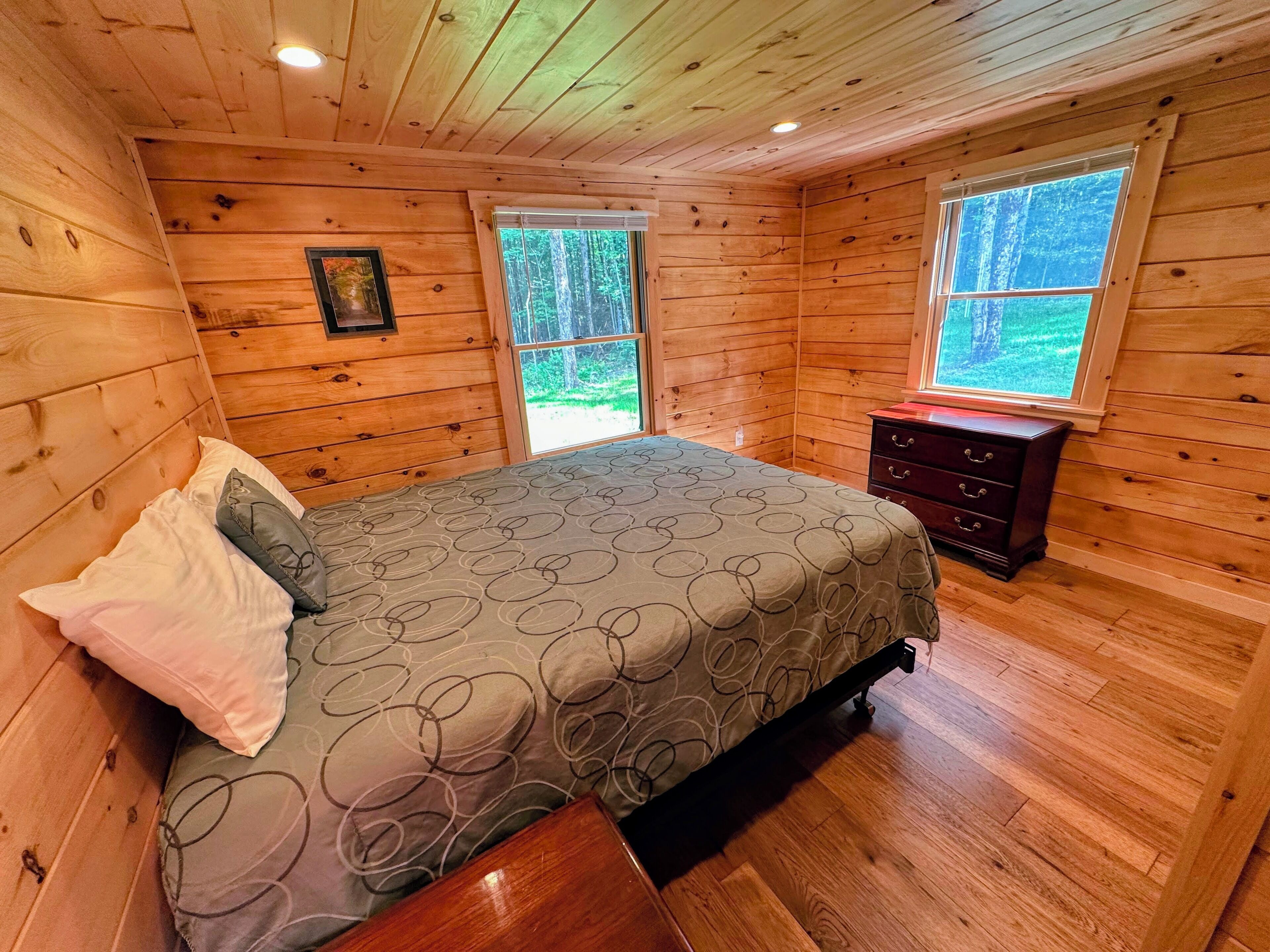 A wooden cabin bedroom with a bed, dresser, and two windows offering a view of the forest.