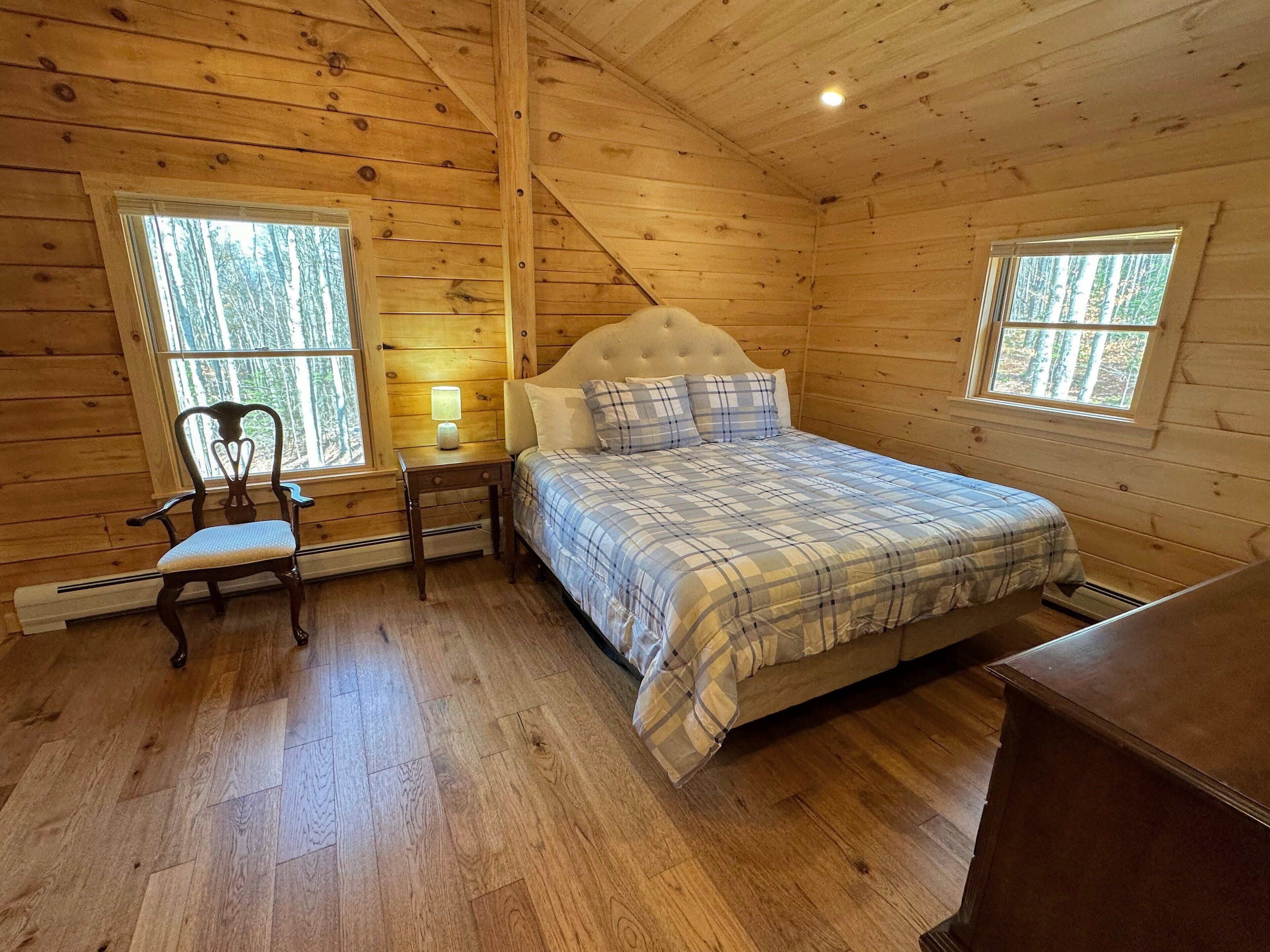A wooden cabin bedroom with a bed, nightstand, chair, and two windows.
