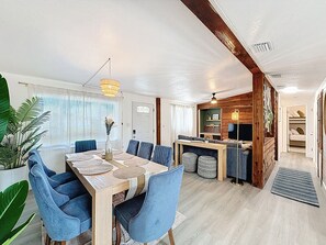 Een ruime eethoek met een houten tafel en blauwe stoelen, een keuken met houten kasten, en een grote plant in de hoek.