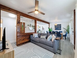 Een moderne woonkamer met een grijze bank, houten details, een plafondventilator en een eethoek op de achtergrond.