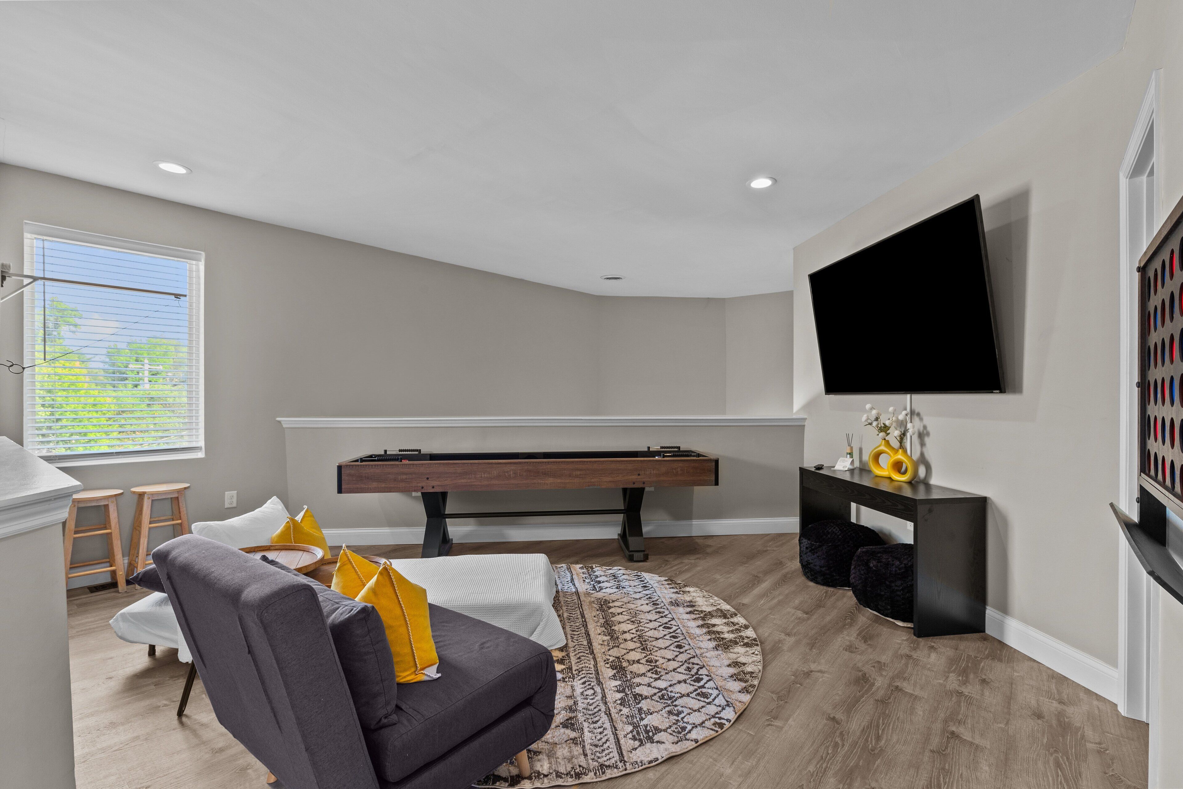 A living room with a pool table, a gray sofa, a round rug, and a flat-screen TV mounted on the wall.