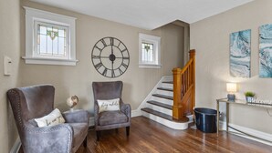 Una sala de estar con una escalera de madera, dos sillones con cojines decorativos, un reloj de pared grande y un letrero de bienvenida.