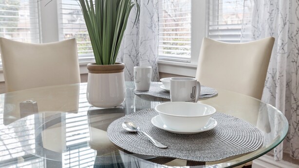 Una mesa de comedor con cubierta de cristal transparente, rodeada por dos sillas, una planta en maceta y un juego de vajilla blanca.