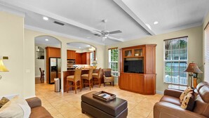 A spacious living area with a sofa, ottoman, and a TV cabinet. There is a dining area with chairs and a table, and a kitchen in the background. - Avenida De Cortez (Siesta Key)