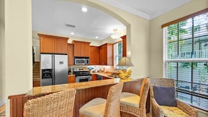 A kitchen with wooden cabinets, a stainless steel refrigerator, a microwave, and a stove. There are wicker chairs around a bar area with a lamp on the counter. - Avenida De Cortez (Siesta Key)
