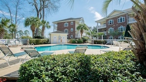 A swimming pool surrounded by lounge chairs, palm trees, and a building with a brick facade. - River Walk 301 (Myrtle Beach)