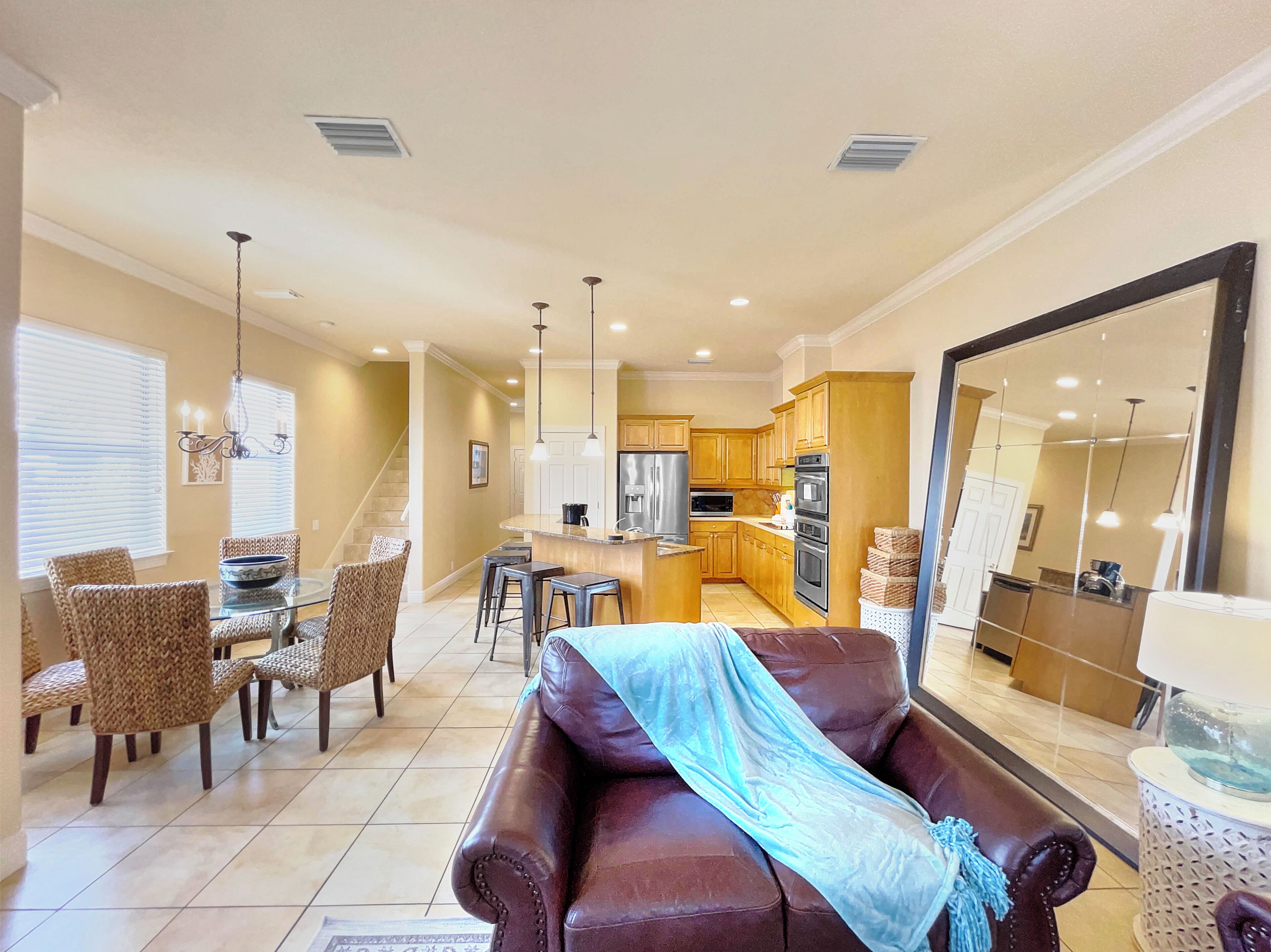 A spacious living room with a leather sofa, a dining area with wicker chairs, a kitchen with wooden cabinets, and a large mirror reflecting the space.