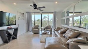 A modern living room with a large TV, leather sofa, and a balcony with curtains. - Sandy Cove - Unit 401 (Siesta Key)
