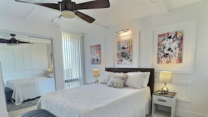 A bedroom with a large bed, two bedside tables with lamps, a ceiling fan, and artwork on the walls. - Sandy Cove - Unit 401 (Siesta Key)