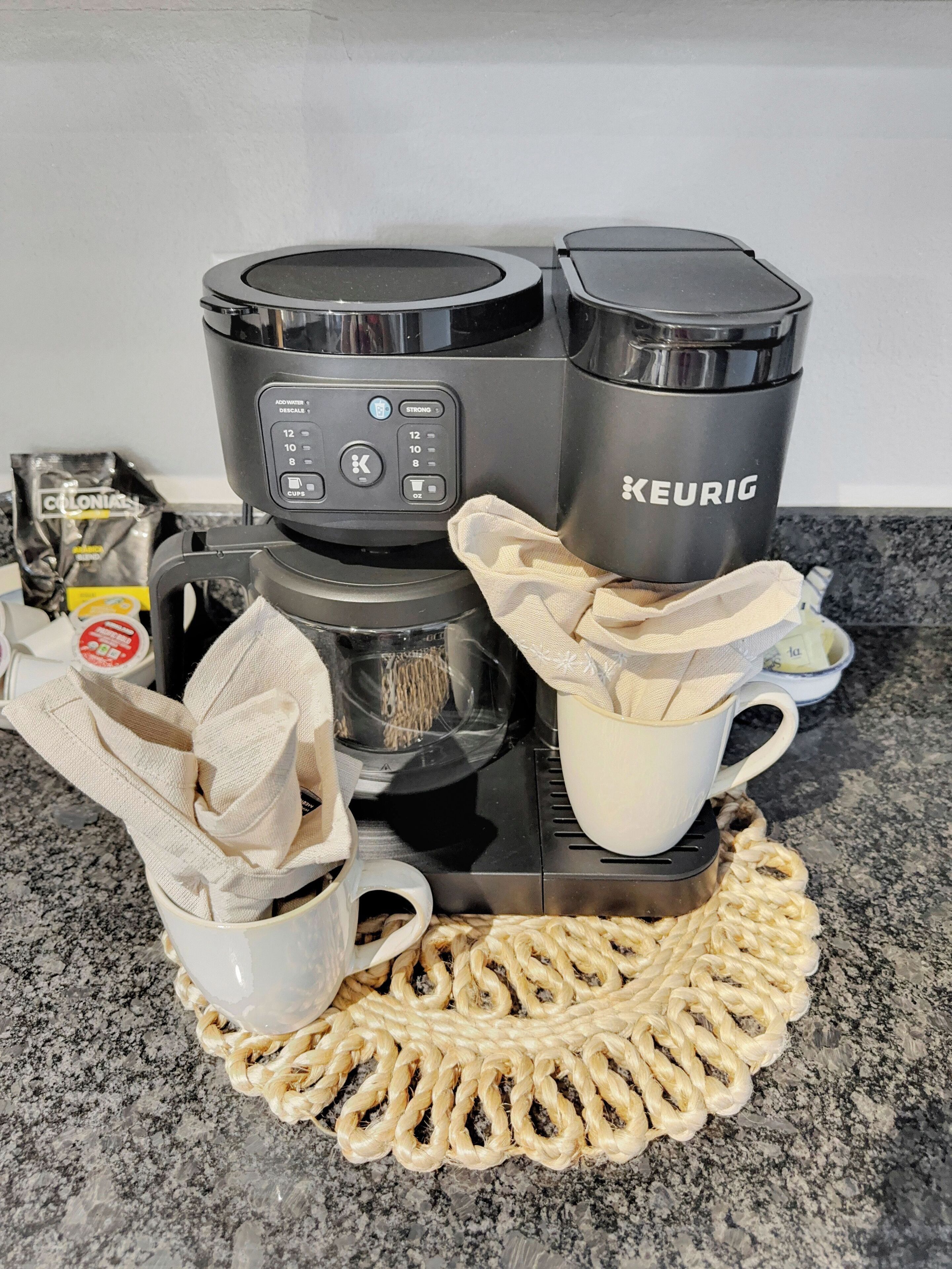 Eine Kaffeemaschine von Keurig, eine Kapsel, eine weiße Tasse und ein Stoffserviettenbild auf einer Granitarbeitsplatte.