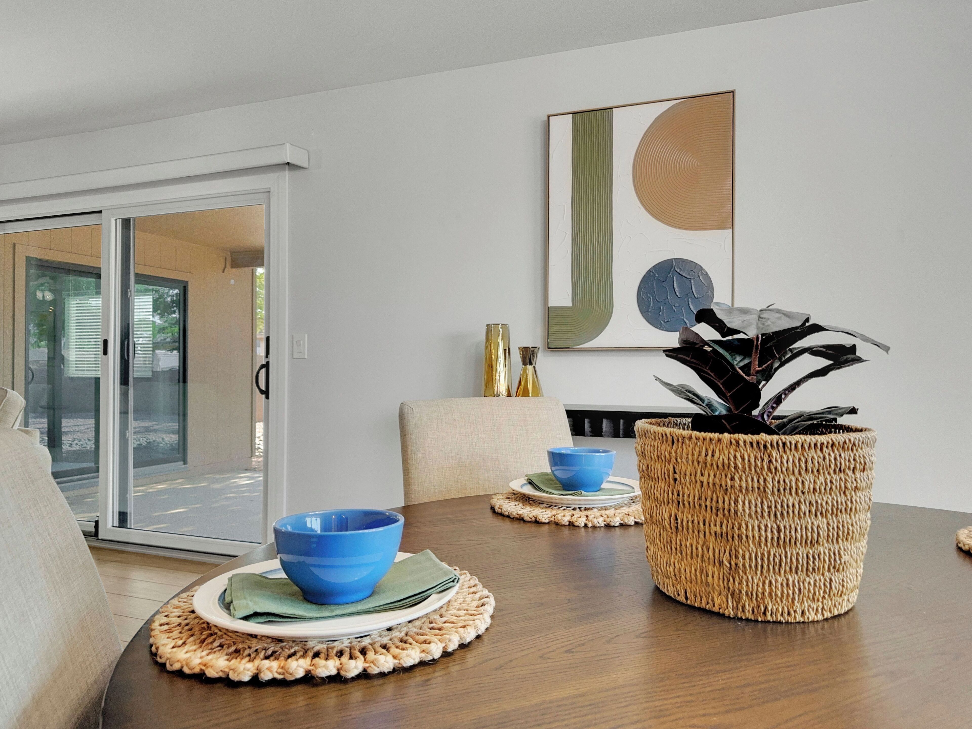 Un comedor con mesa de madera, cuencos azules, una cesta tejida y un cuadro abstracto colgado en la pared.