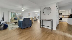Amplia sala de estar con sofá, sillón y mesa de centro, comedor con sillas y cocina con gabinetes y electrodomésticos blancos.