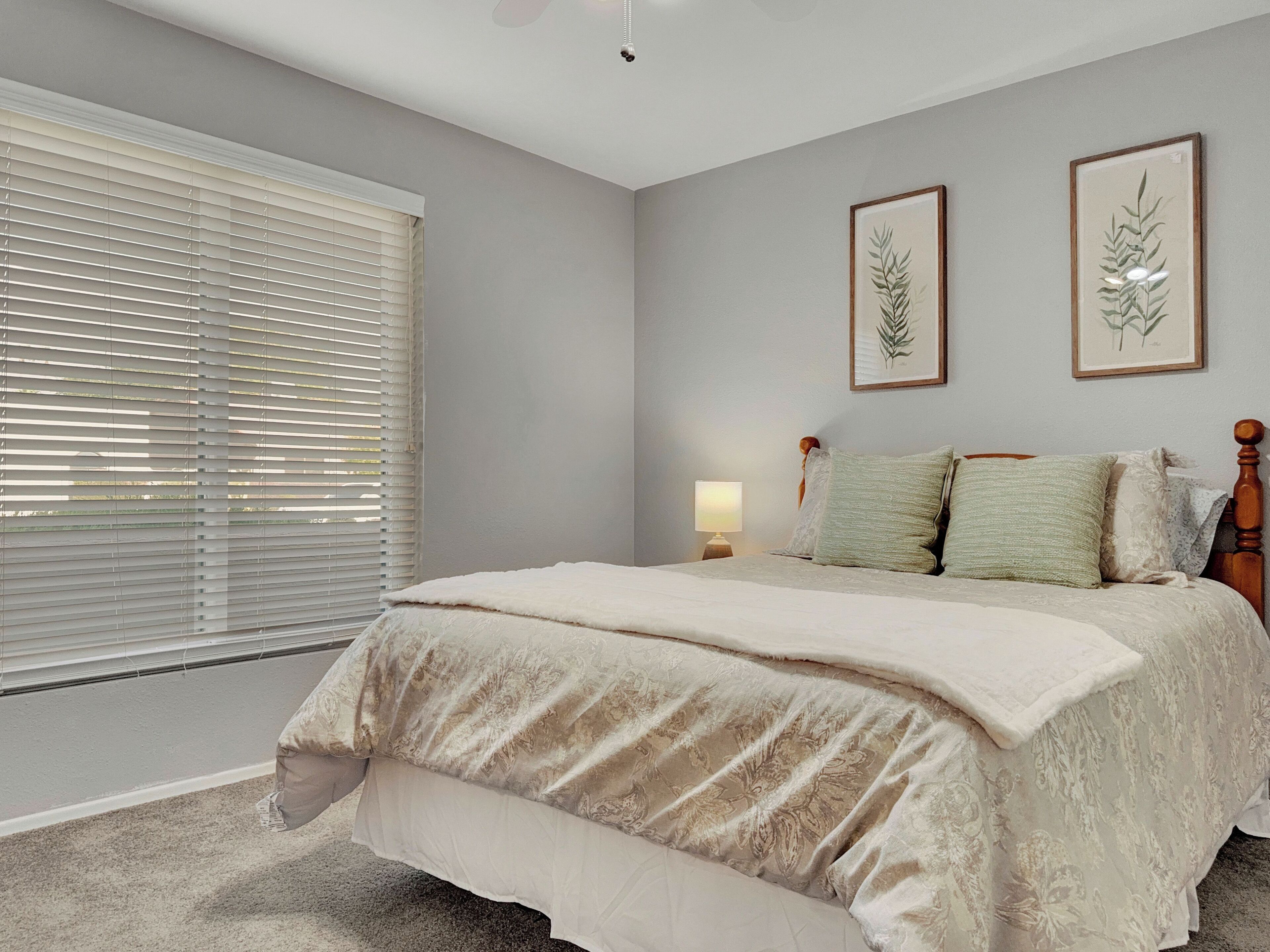 A bedroom with a large bed, a nightstand, a lamp, and two framed botanical prints on the wall.