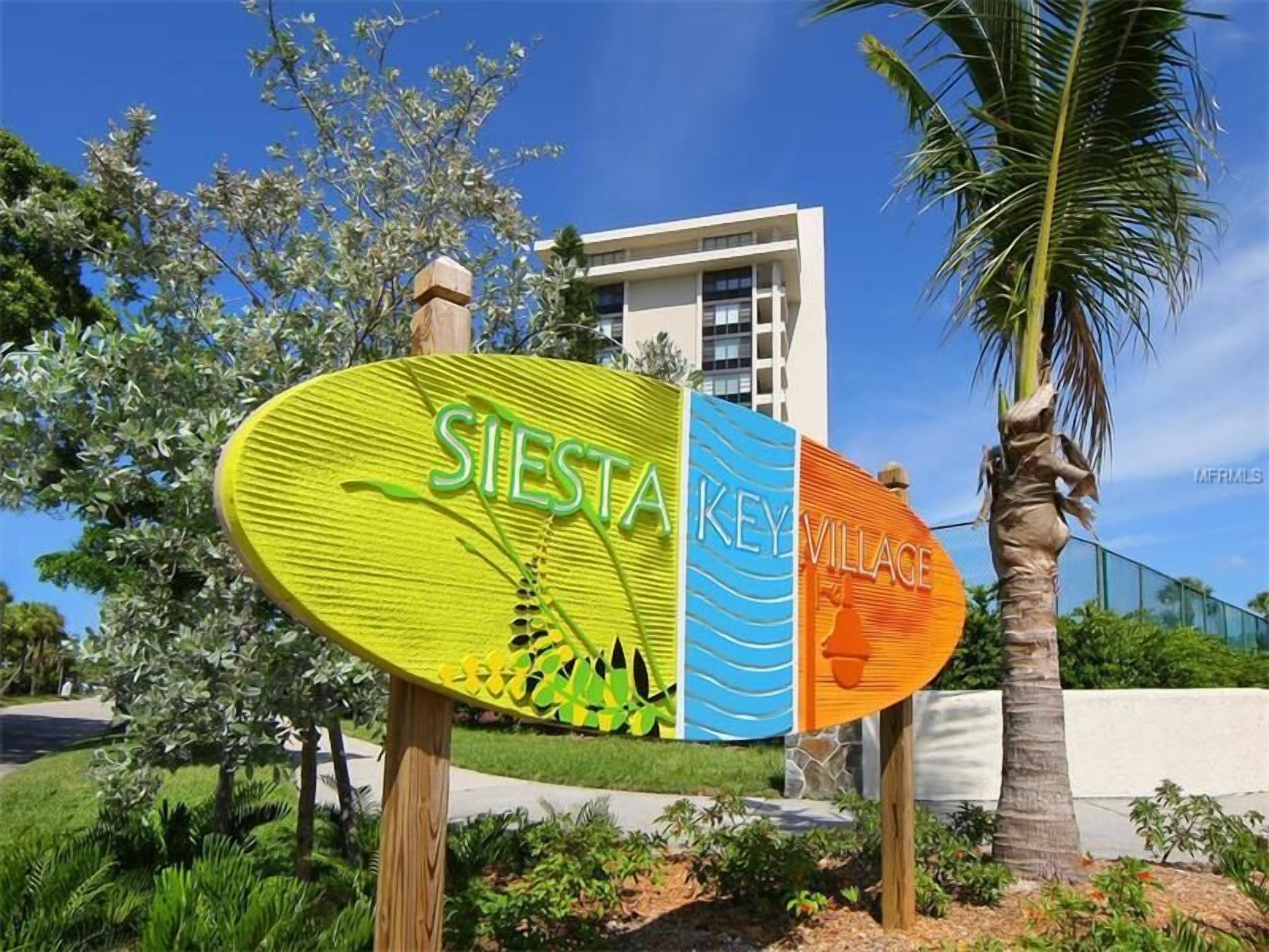 Um letreiro com a inscrição 'SIESTA KEY VILLAGE', rodeado por vegetação e uma palmeira.