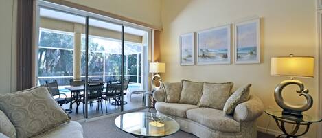 A living room with a glass coffee table, two sofas, and a dining area with chairs and a table. There are three framed pictures on the wall and a lamp on a side table.