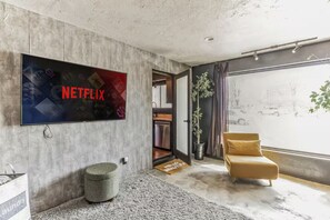 A modern living room with a large flat-screen TV, a grey ottoman, a yellow sofa, and a view of the outdoors through a large window. - Walkable~ Bikes~ Firepits~ Petsok~ Bbq~ Dining (St. George)