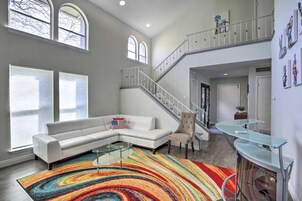 Uma sala de estar espaçosa com um sofá seccional branco, um tapete colorido, uma mesa de centro de vidro e uma escada que leva a um andar superior.